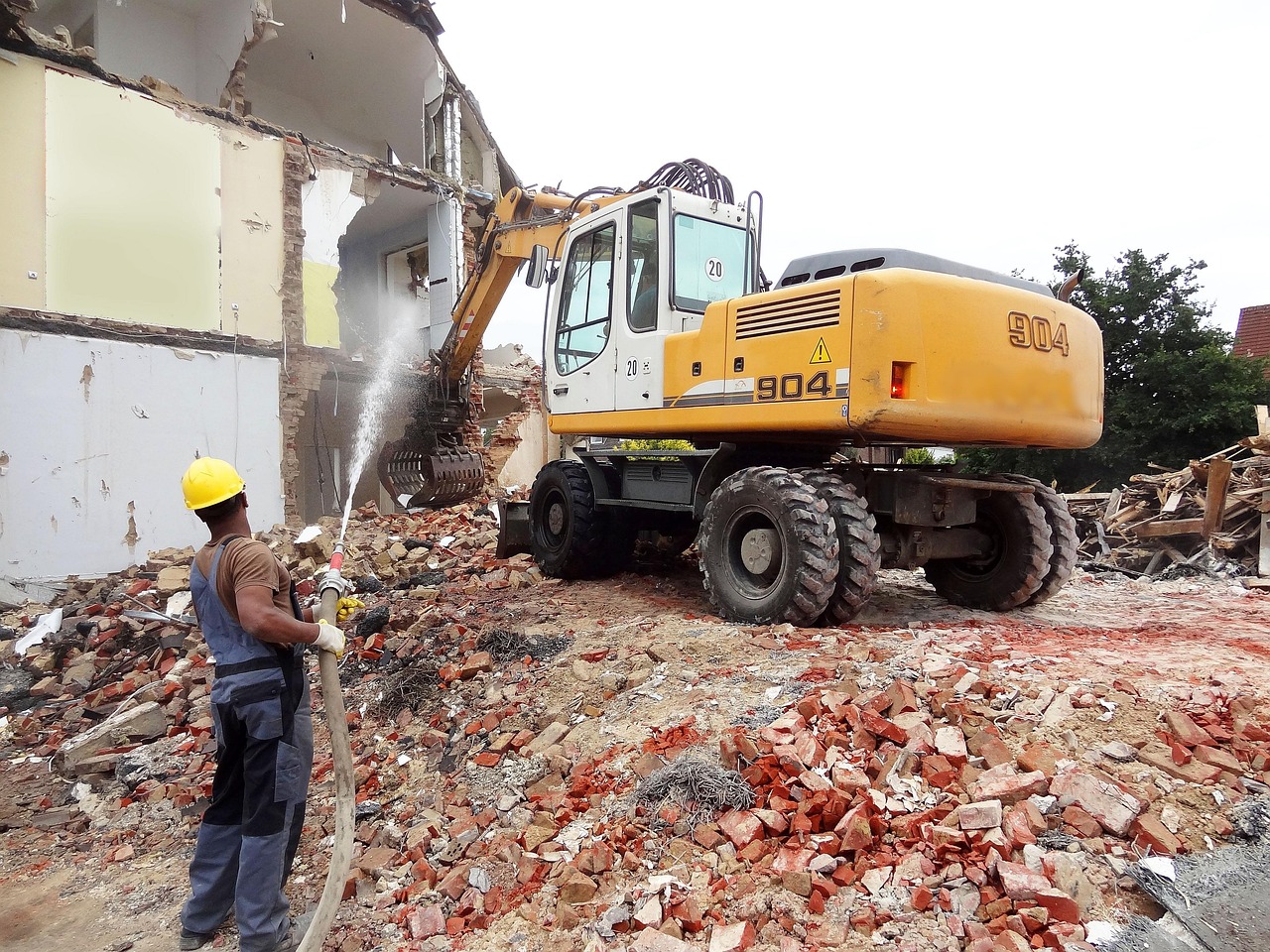Travaux de démolition avec pelleteuse détruisant un bâtiment et ouvrier arrosant pour limiter la poussière.